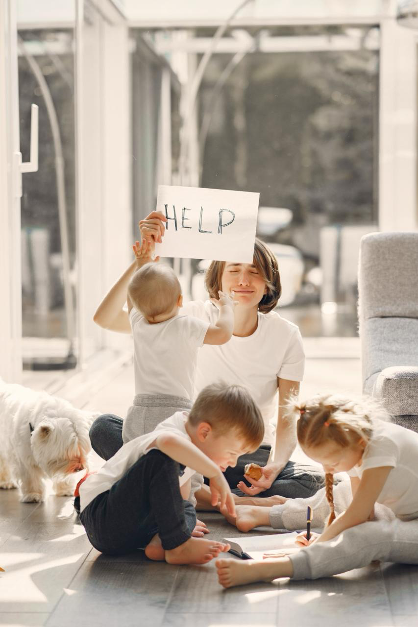 Un parent entouré de trois enfants tient une pancarte HELP dans le salon familial